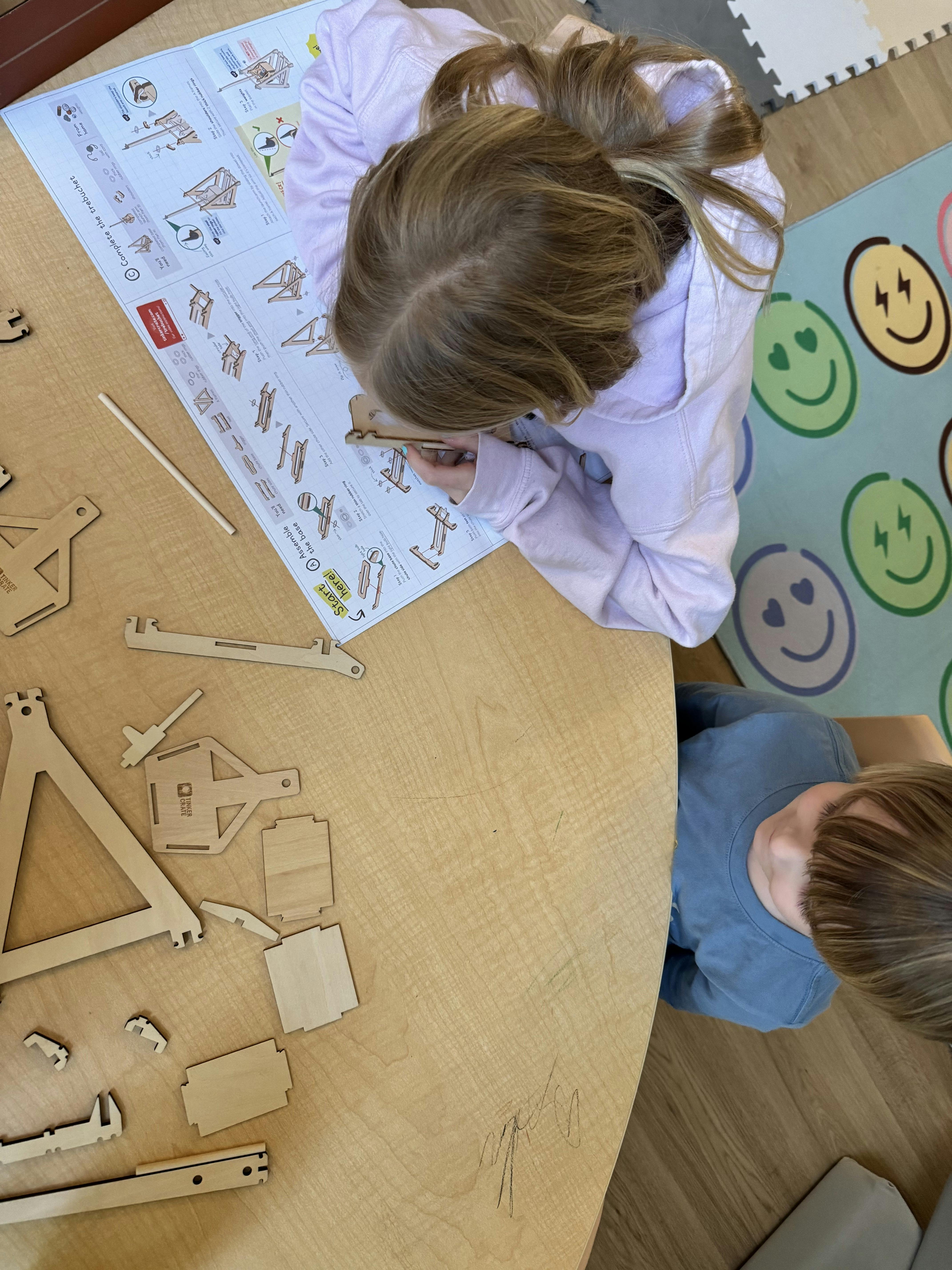 Child assembling a wooden trebuchet from a kit, with another child nearby