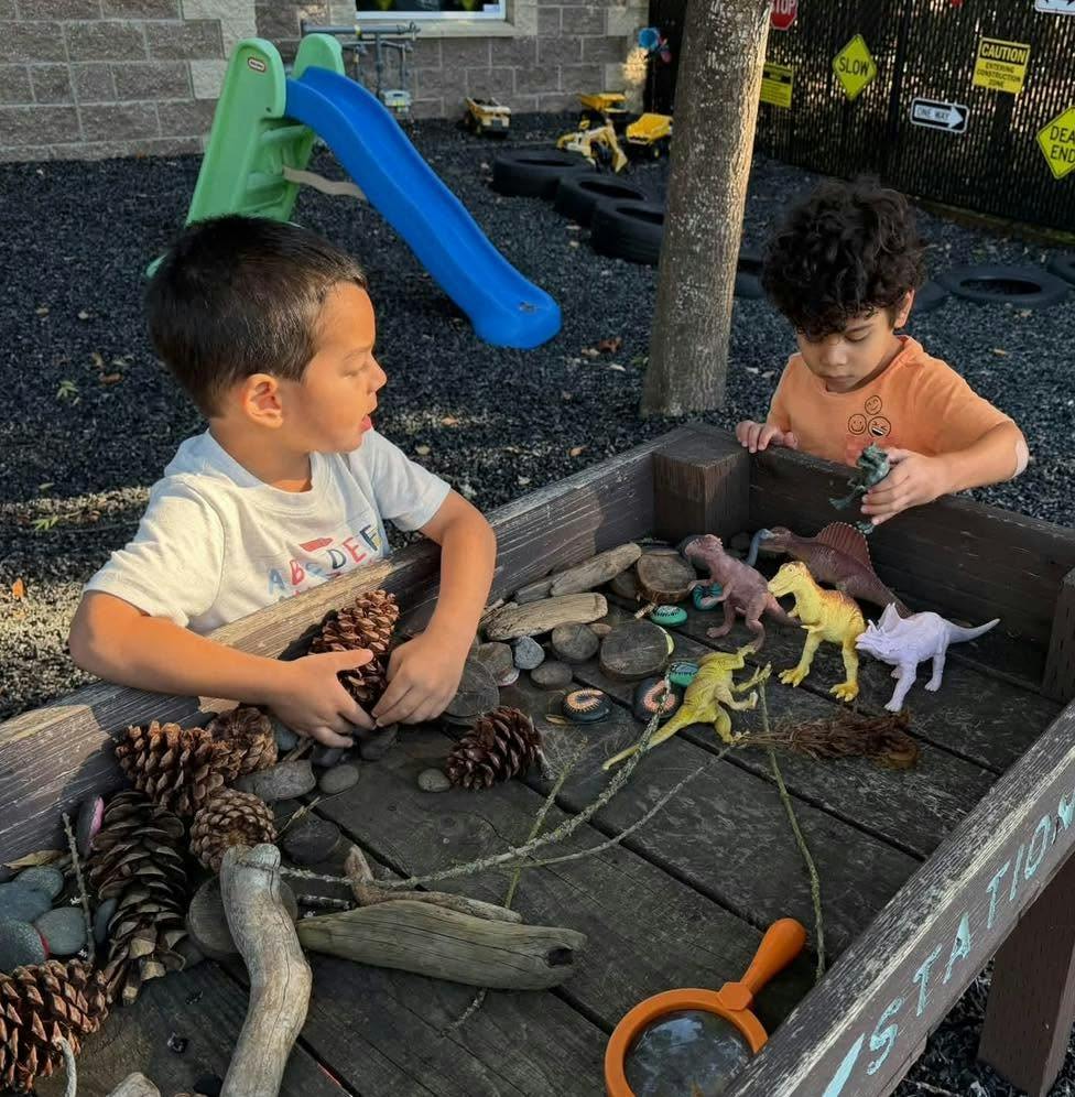 Two children playing with dinosaur figurines in an outdoor sensory bin