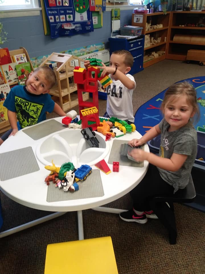 Three children playing with building blocks and toys at a table in a classroom