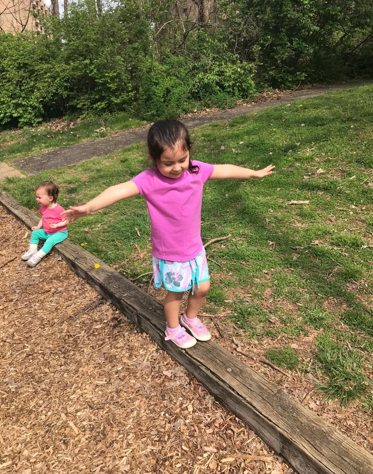 Child balancing on a wooden beam with arms outstretched, another child seated nearby