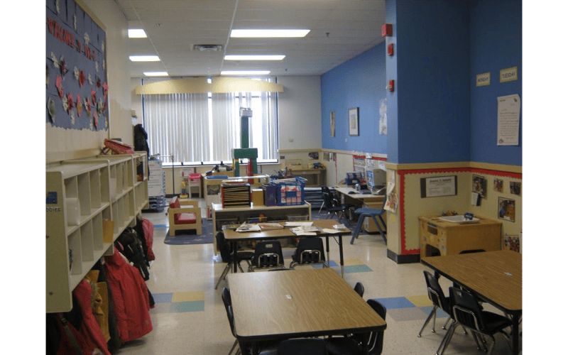 Empty classroom with small tables and chairs, cubbies with coats, and play areas