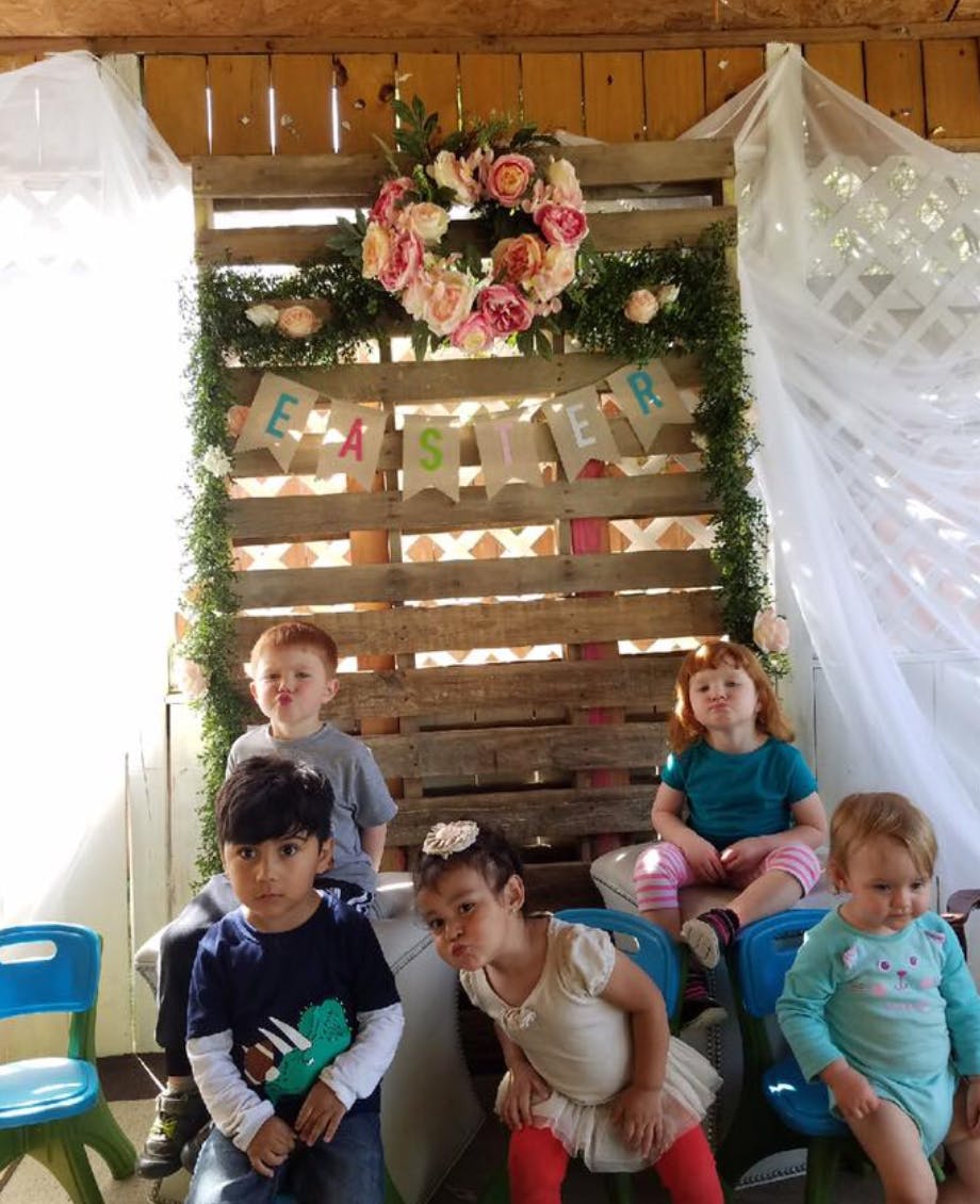 Five children sitting in front of a wooden pallet decorated with a floral wreath and "EASTER" banner