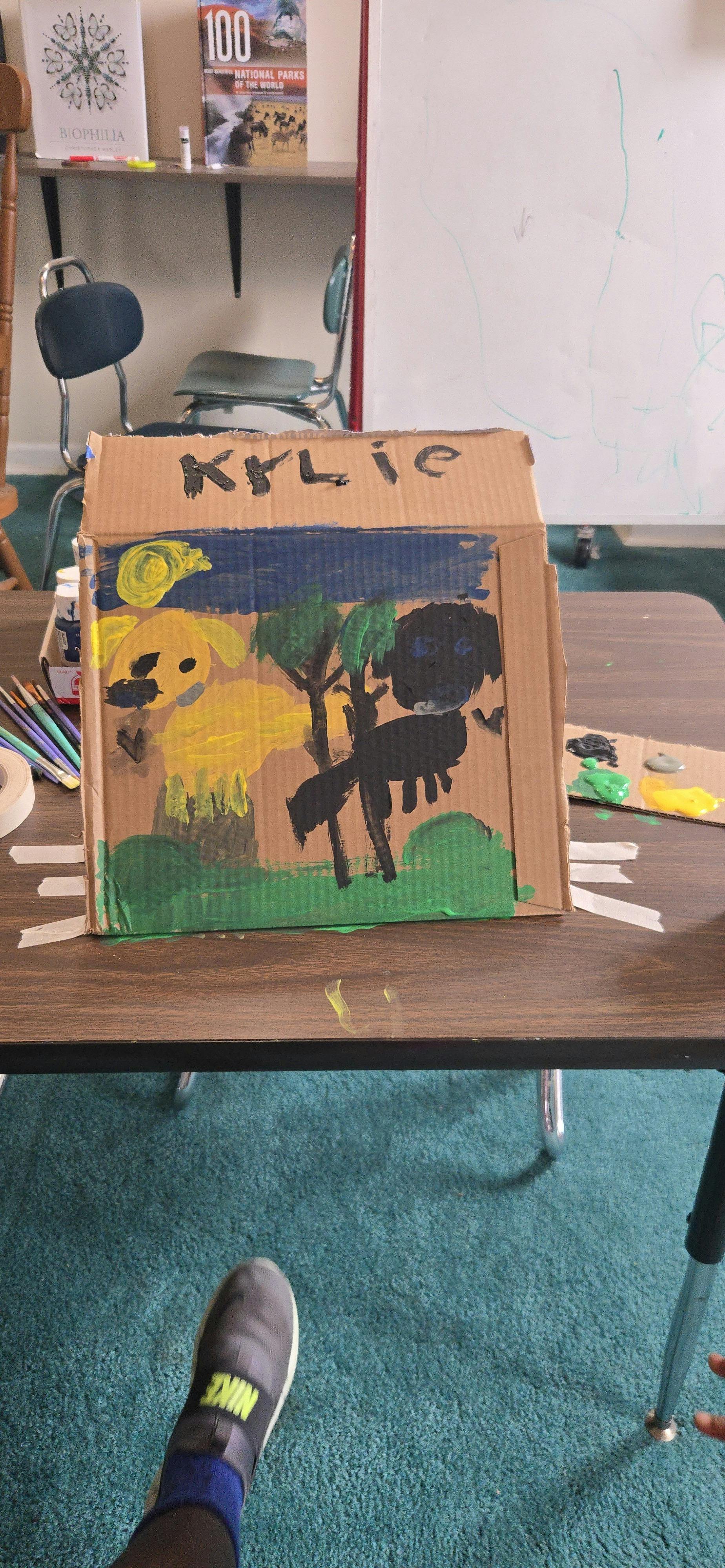 Child painted artwork on cardboard featuring two animals and trees labeled Kylie