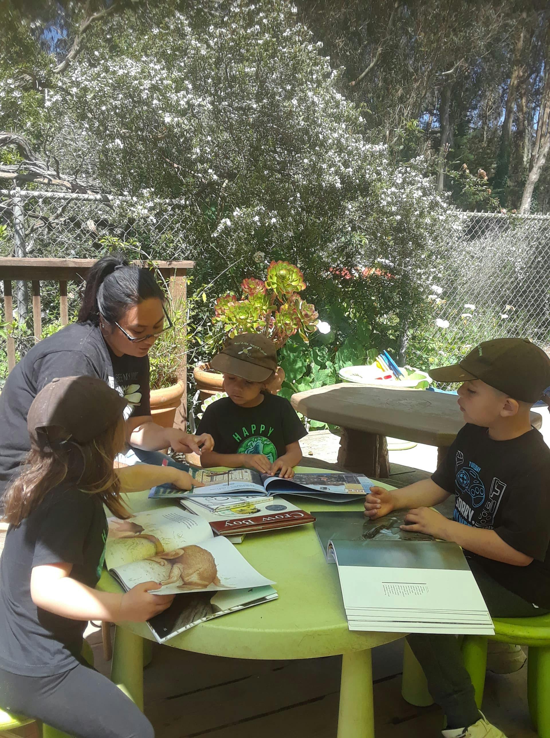 Person and three children reading books at a table outdoors