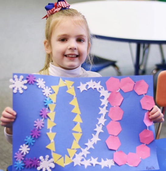 Child holding up a blue poster board with colorful shapes forming "20"