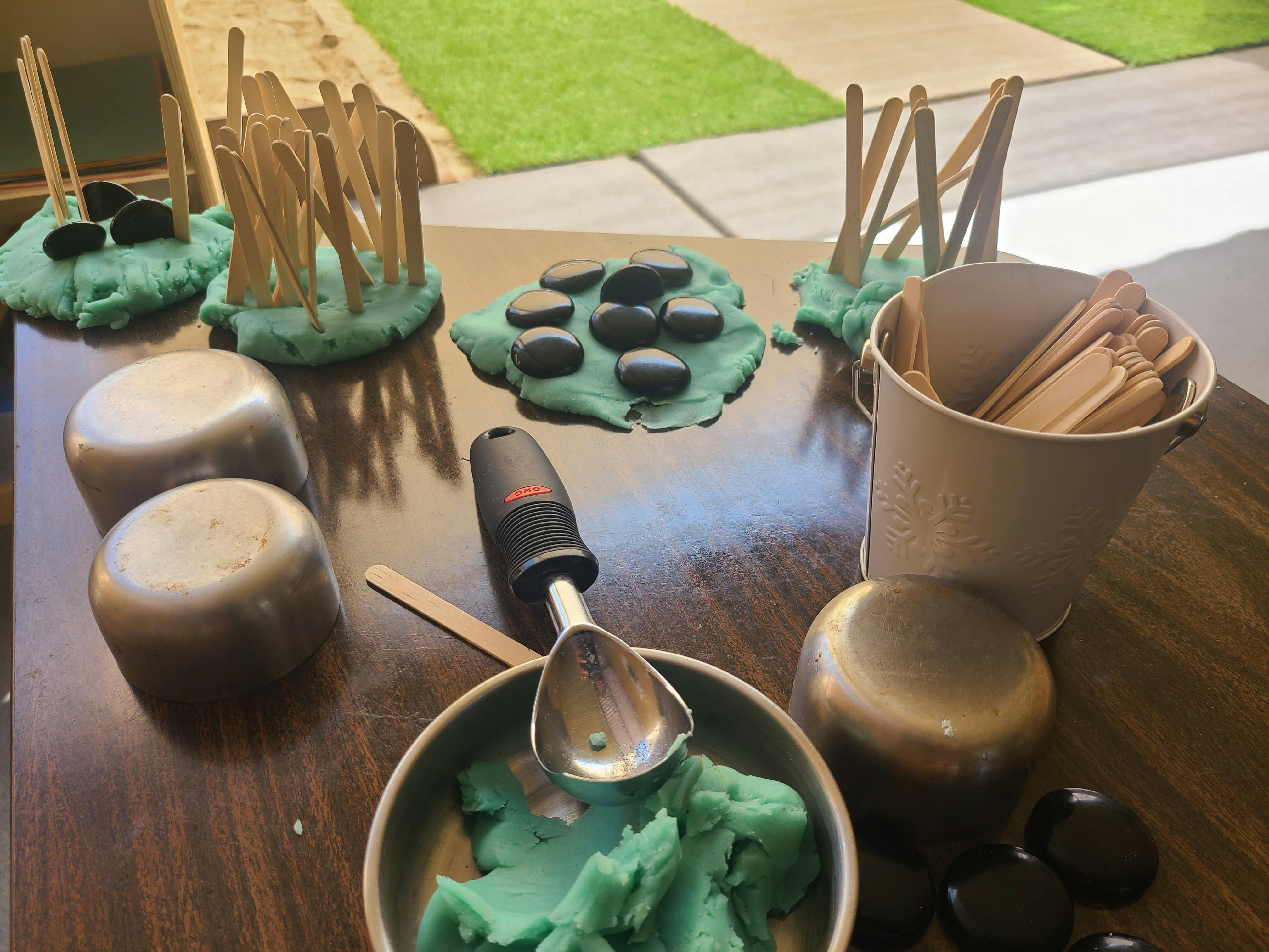 Green playdough with sticks and black stones, metal containers and wooden accessories