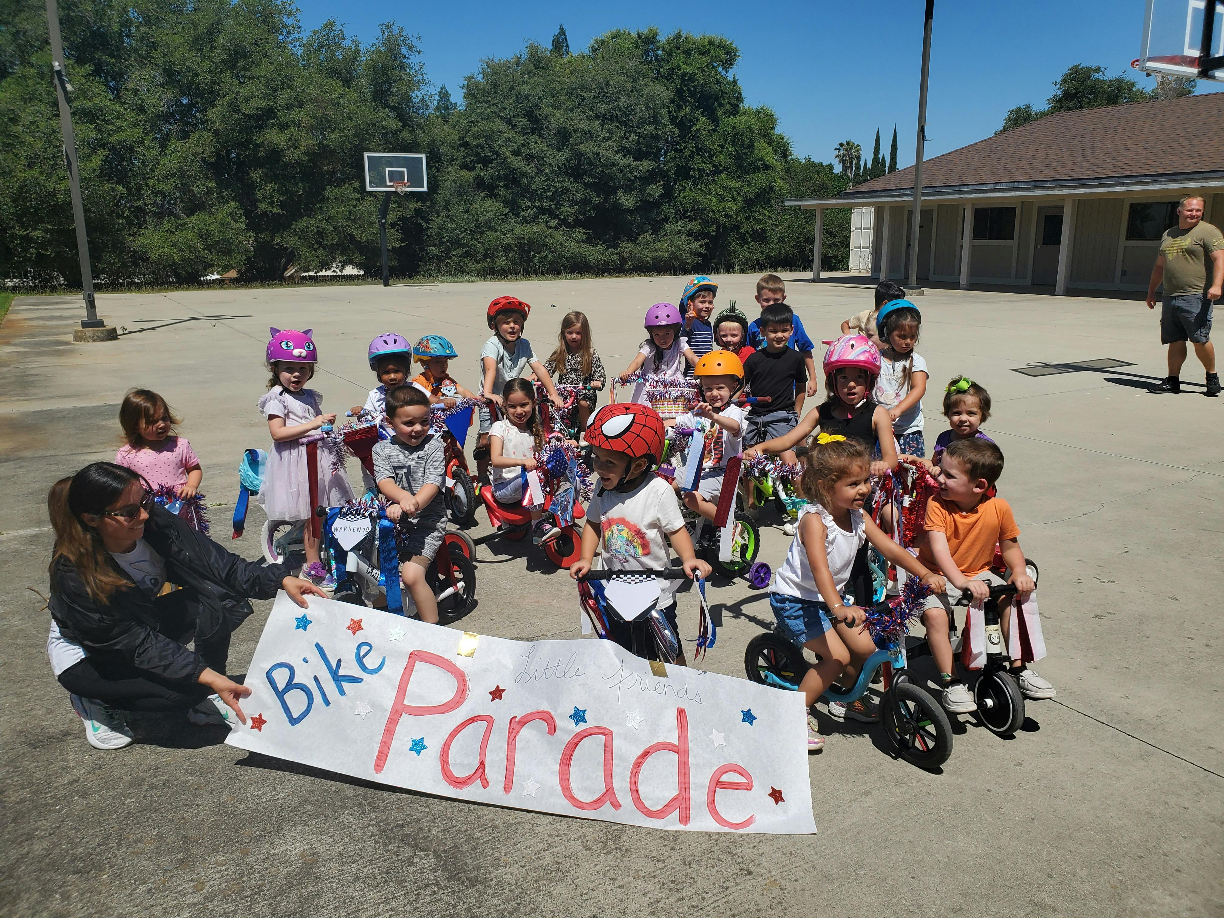 Person holding a "Bike Parade" sign with children on bikes and scooters