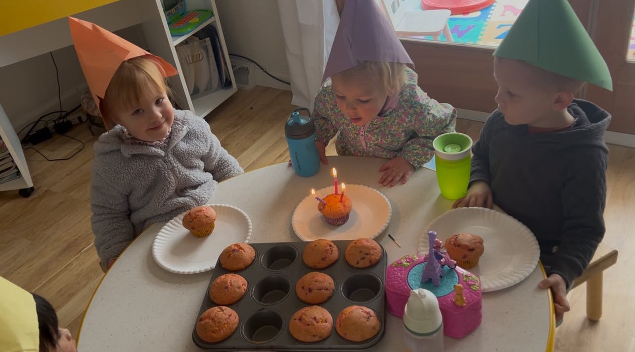 Three children wearing party hats at a table with cupcakes and a lit candle