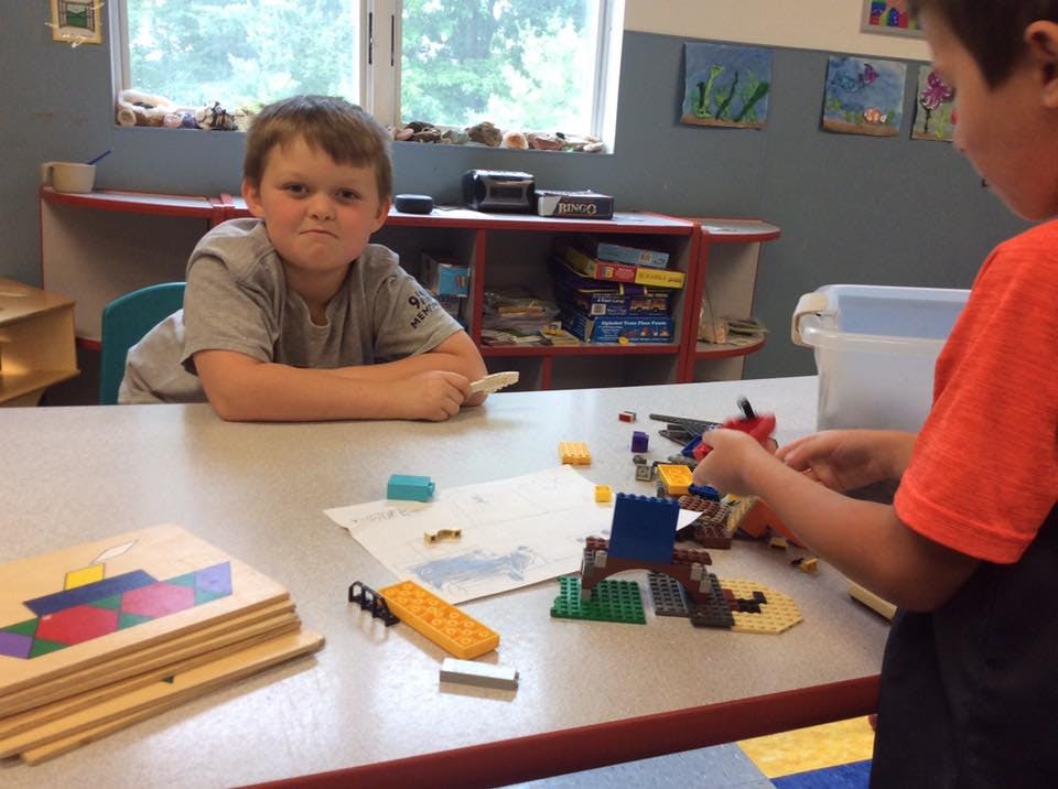 Child sitting at a table while another child plays with building blocks