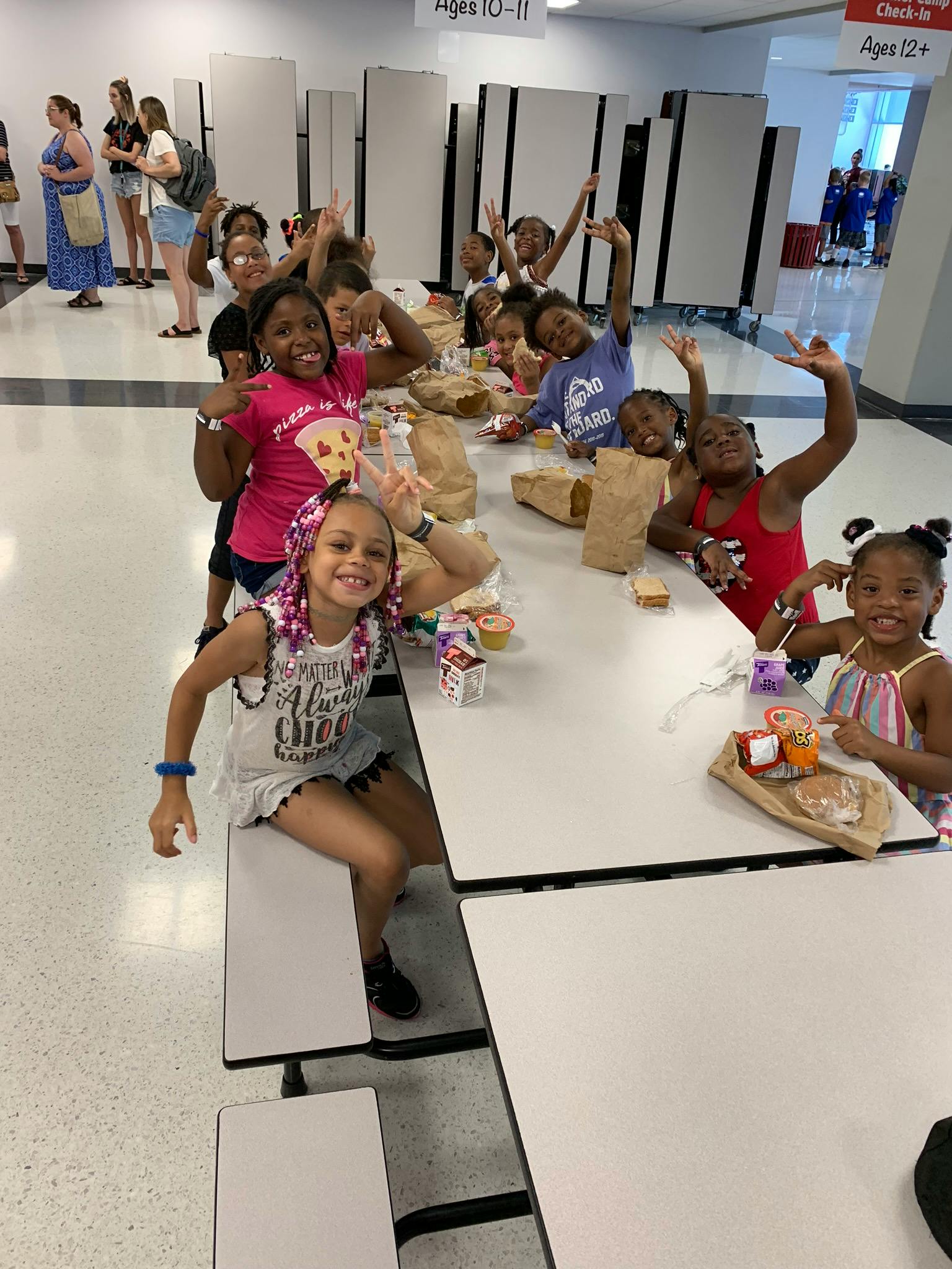 Children sitting at tables with food, some waving