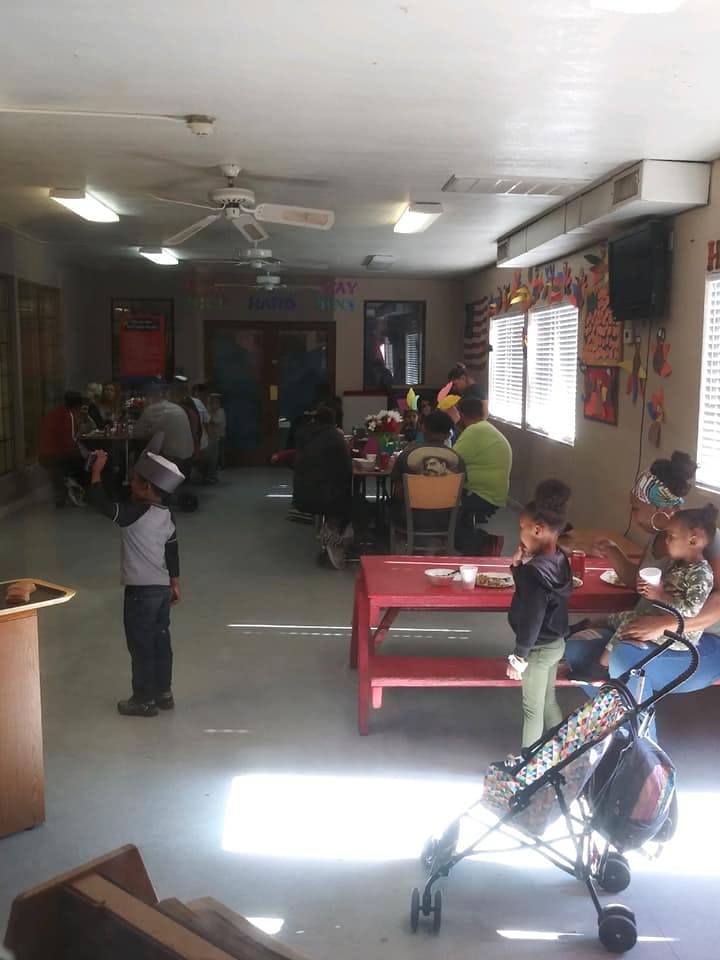 People gathered in a room, some seated at tables, a child standing in foreground