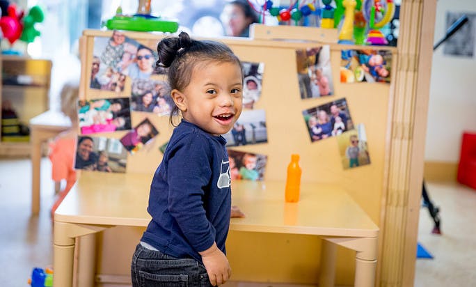 Child smiling at the camera, standing next to a table with photos in the background