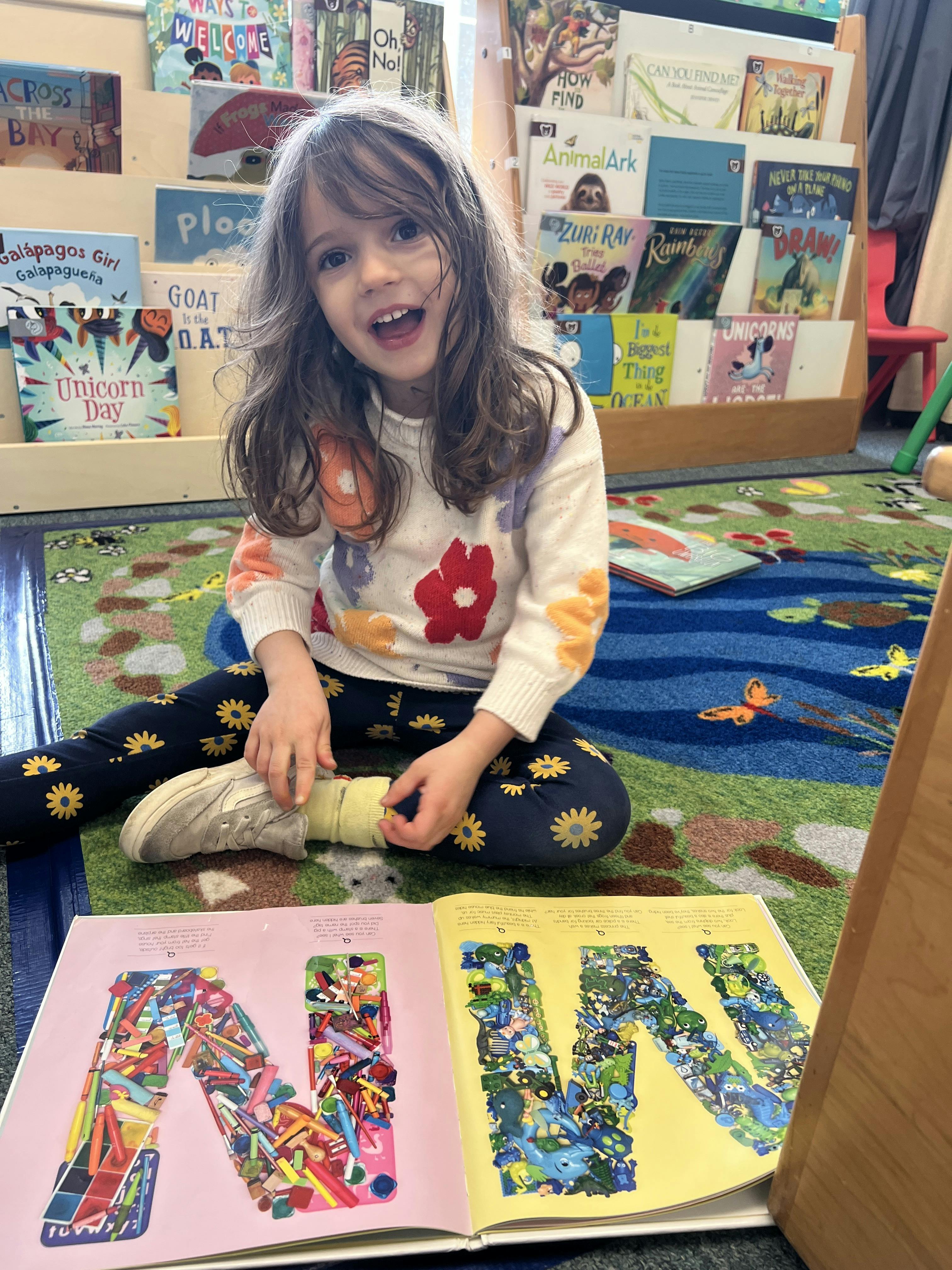 Child sitting on a rug with an open book, smiling at the camera