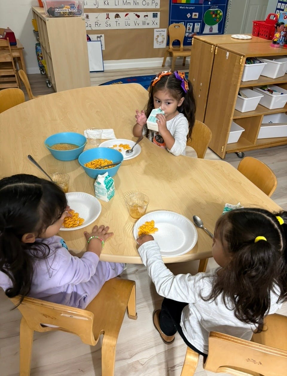Three children sitting at a table eating cereal in a classroom