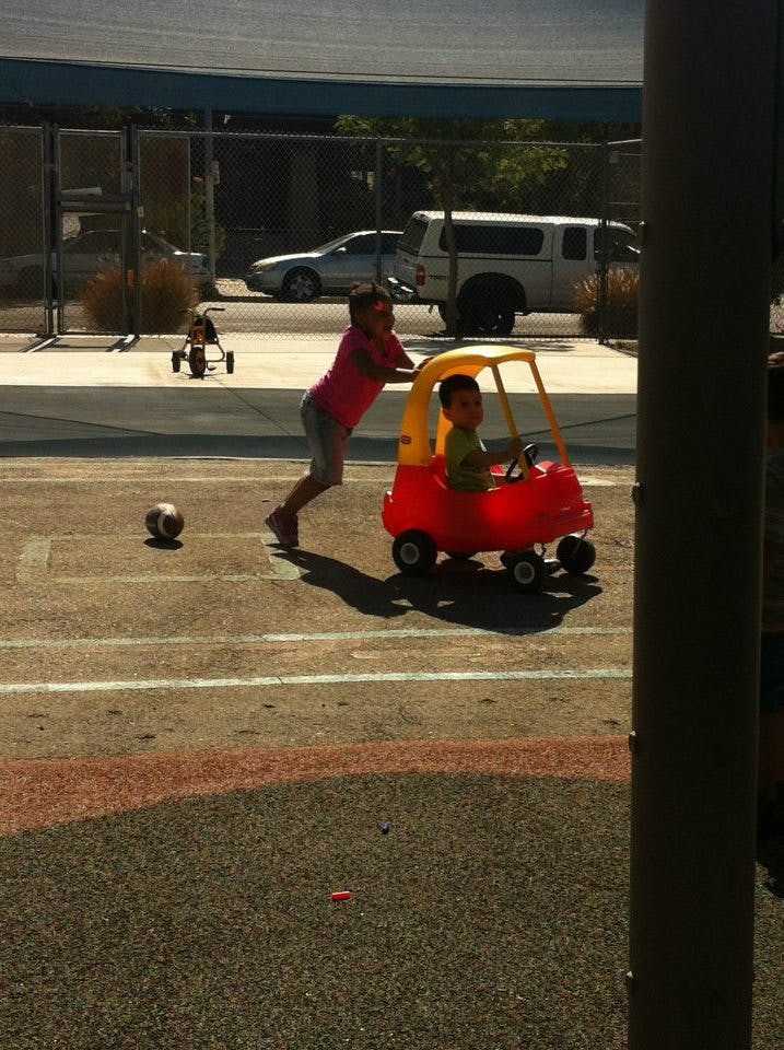 Child pushing another child in a toy car on a paved surface
