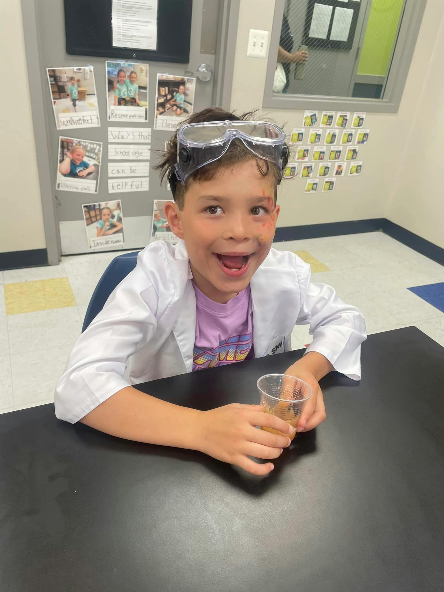 Child in lab coat and safety goggles holding a cup