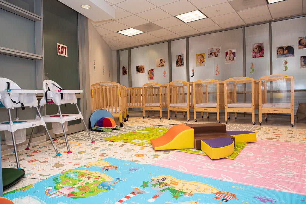 Cribs and high chairs in a daycare room with colorful floor mats