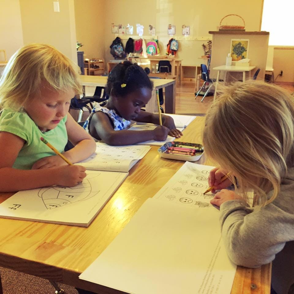 Three children drawing and writing at a table in a classroom