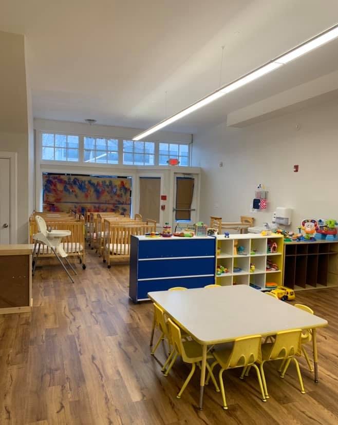 Empty daycare room with cribs, small tables, and storage shelves