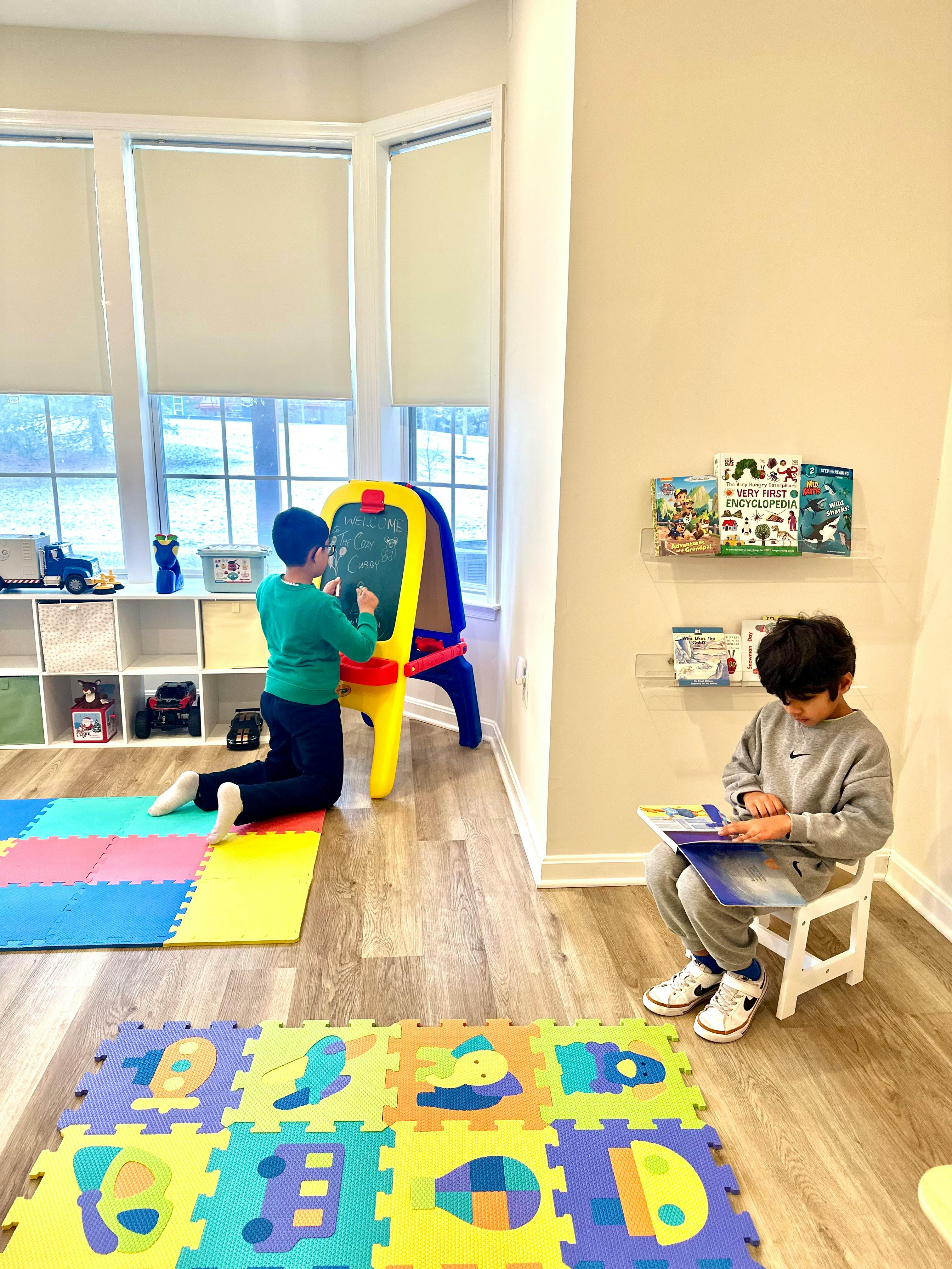 Two children in a room, one drawing on an easel, one reading a book
