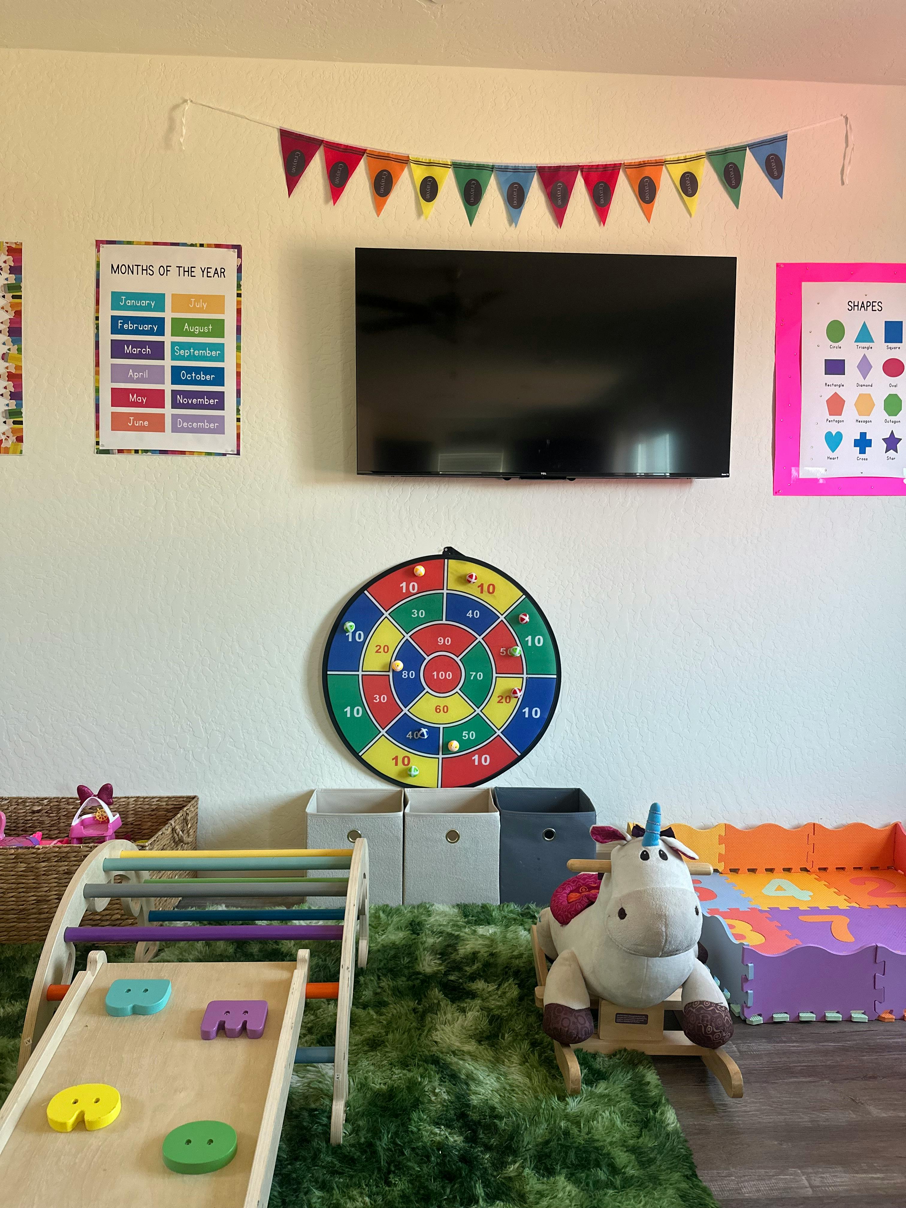 Playroom wall with a television, dartboard, shape chart, and unicorn rocker