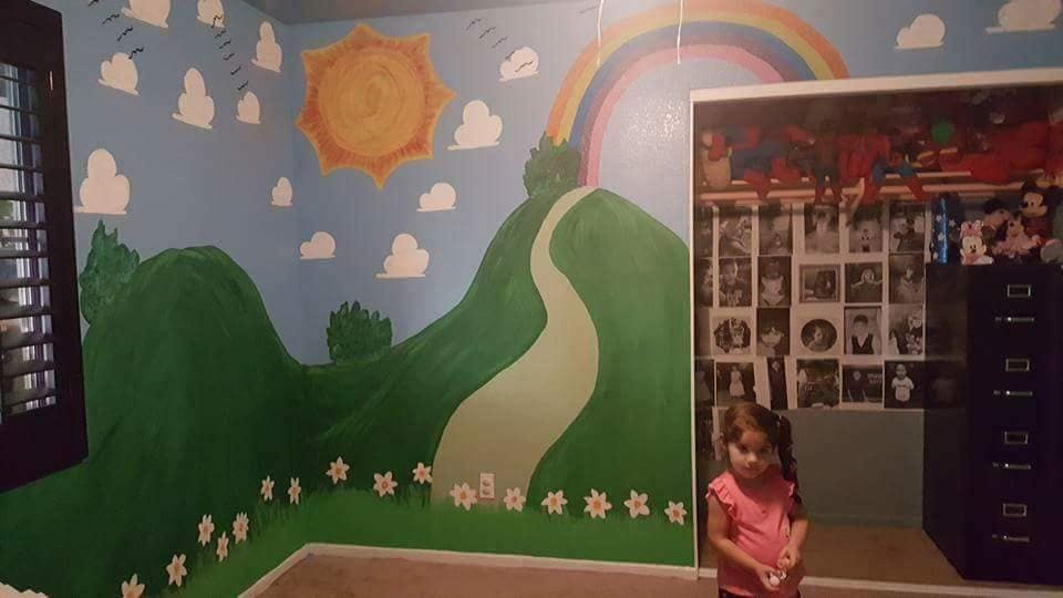 Child standing in a room with a painted mural of hills, sun, and rainbow