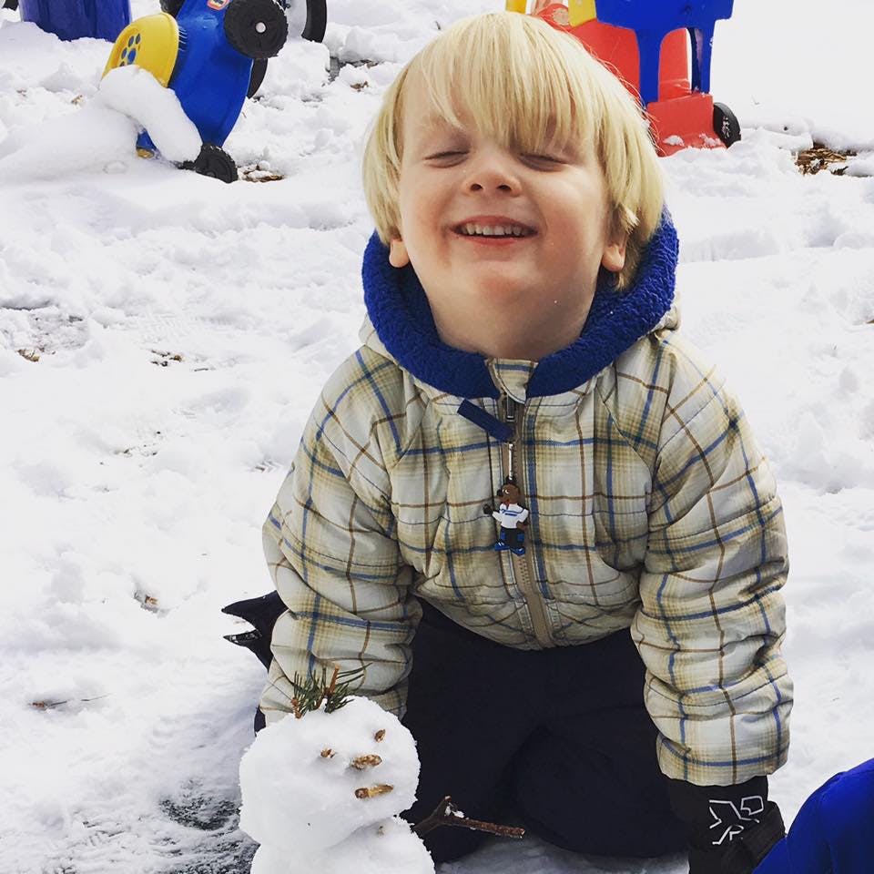 Child in plaid jacket kneeling in snow next to small snowman
