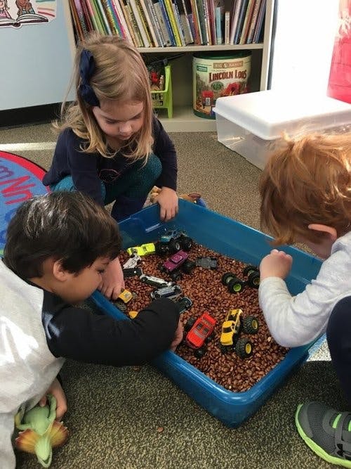 Three children playing with toy cars in a bin of beans