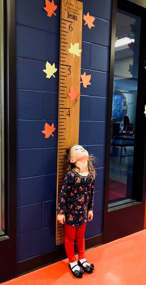 Child standing next to a wooden growth chart with "Jesus" written on it