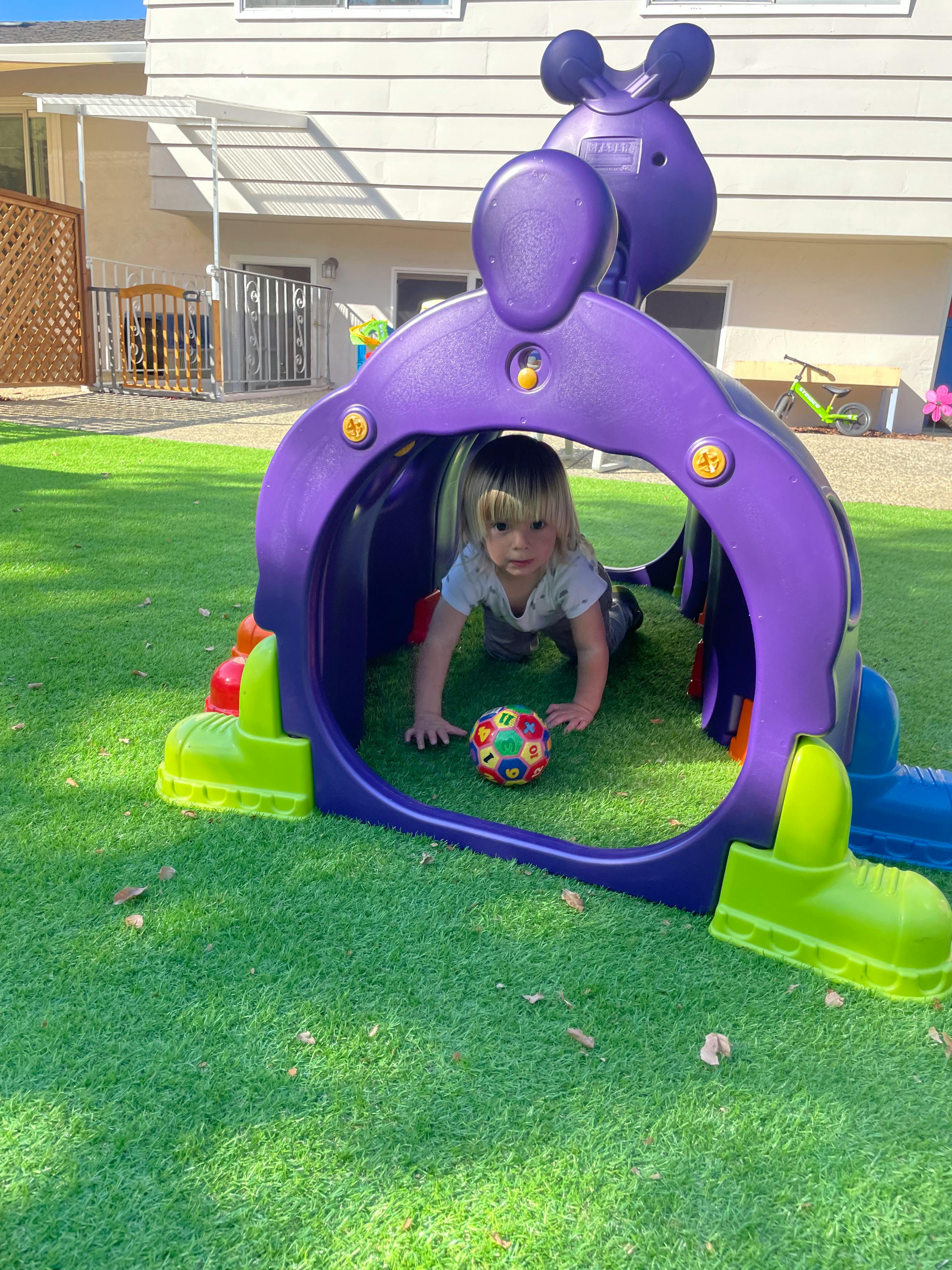 Child crawling through a purple play tunnel with a ball