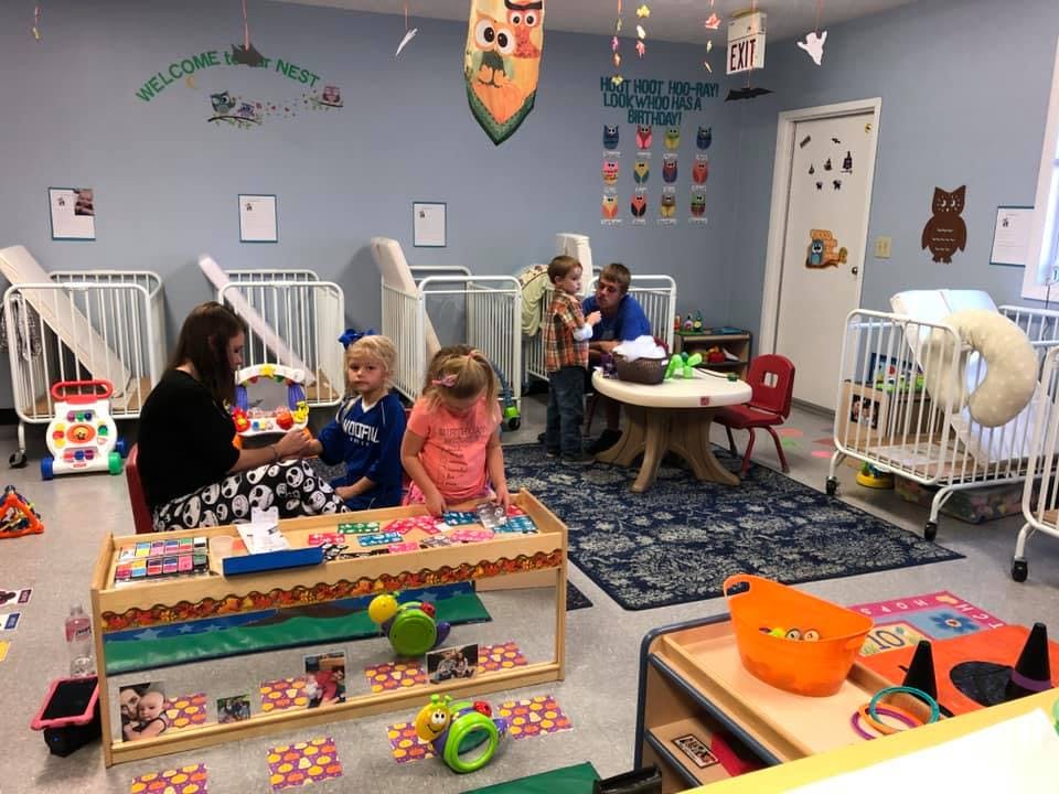 Adults and children in a daycare room with cribs and toys