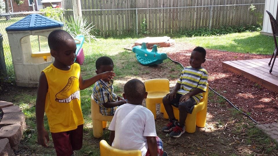 Four children playing outdoors with small chairs and a playhouse