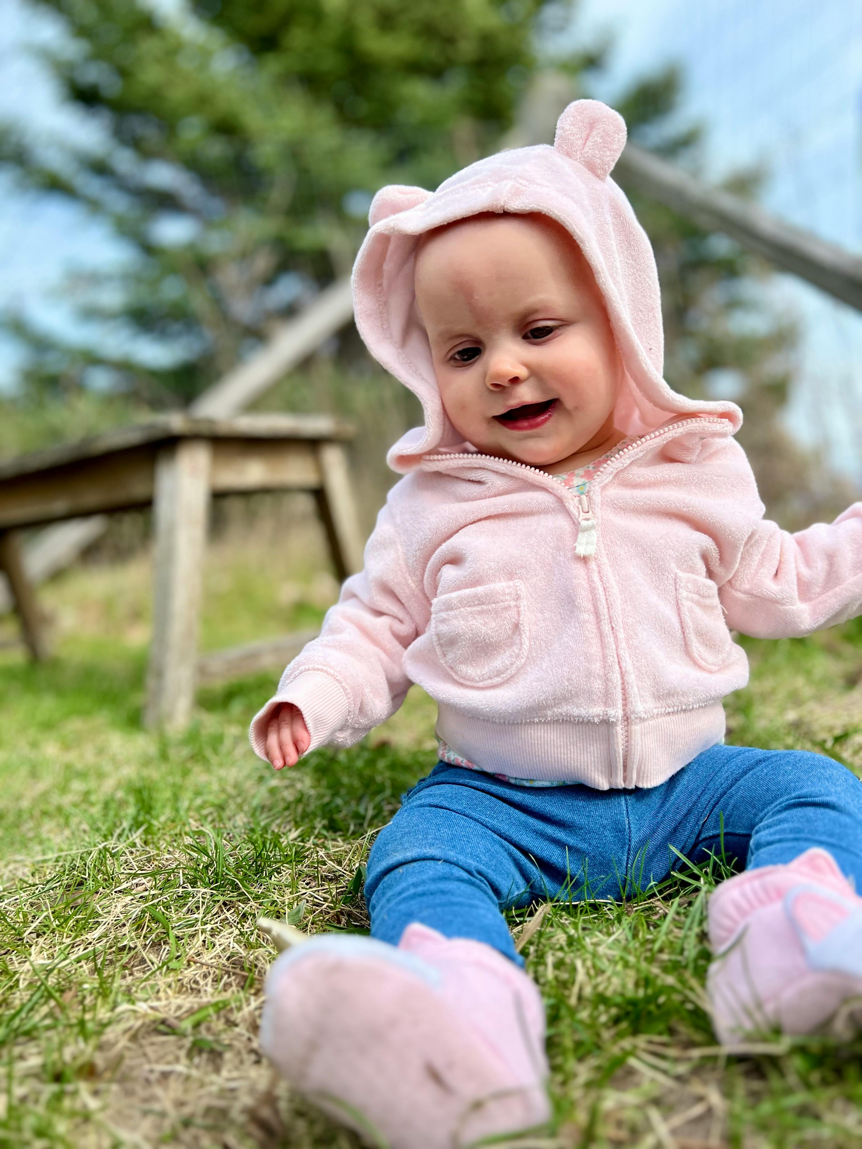 Child in a pink hooded jacket and blue pants sitting on grass