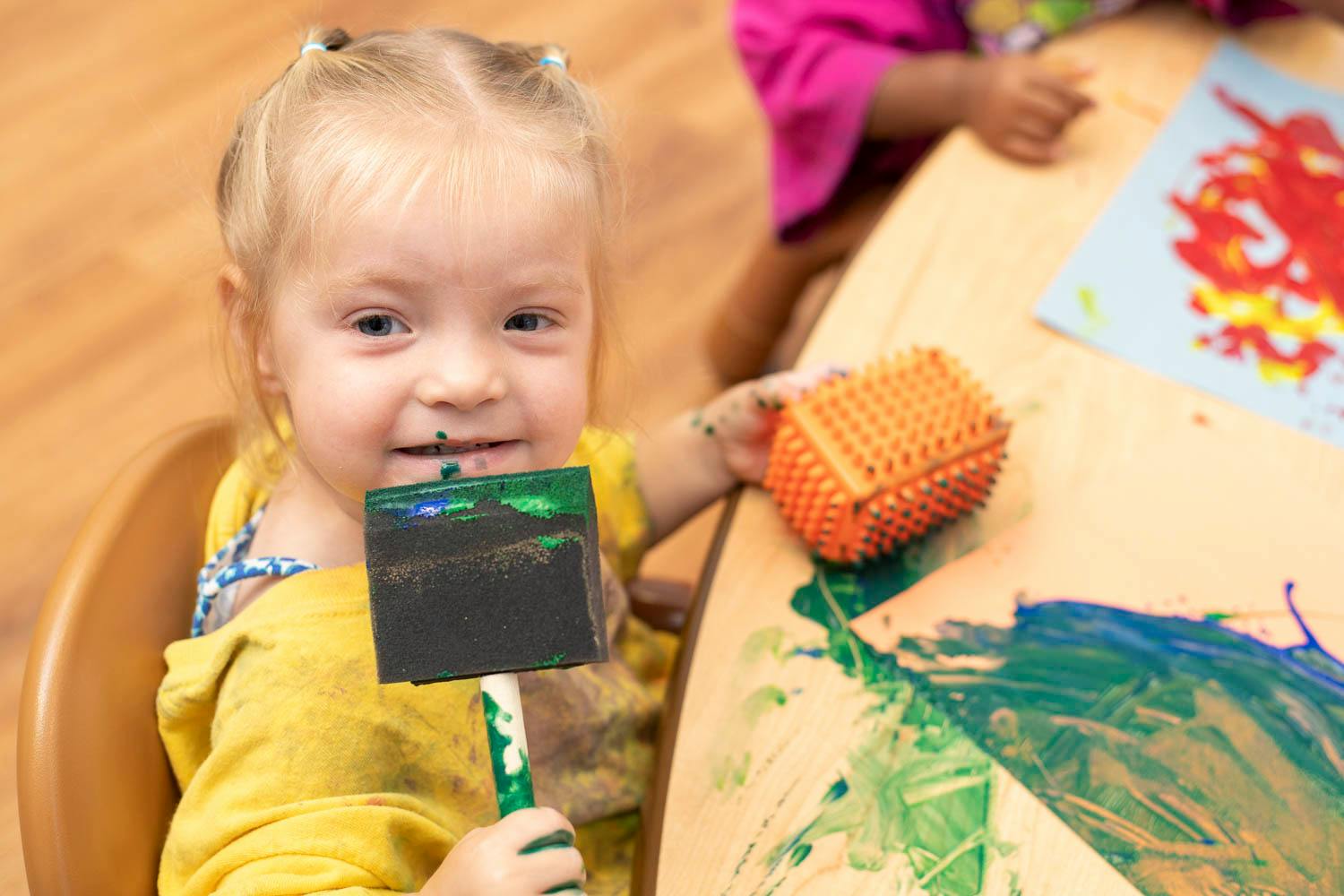 Child holding a paint brush and a textured roller at a table with paint