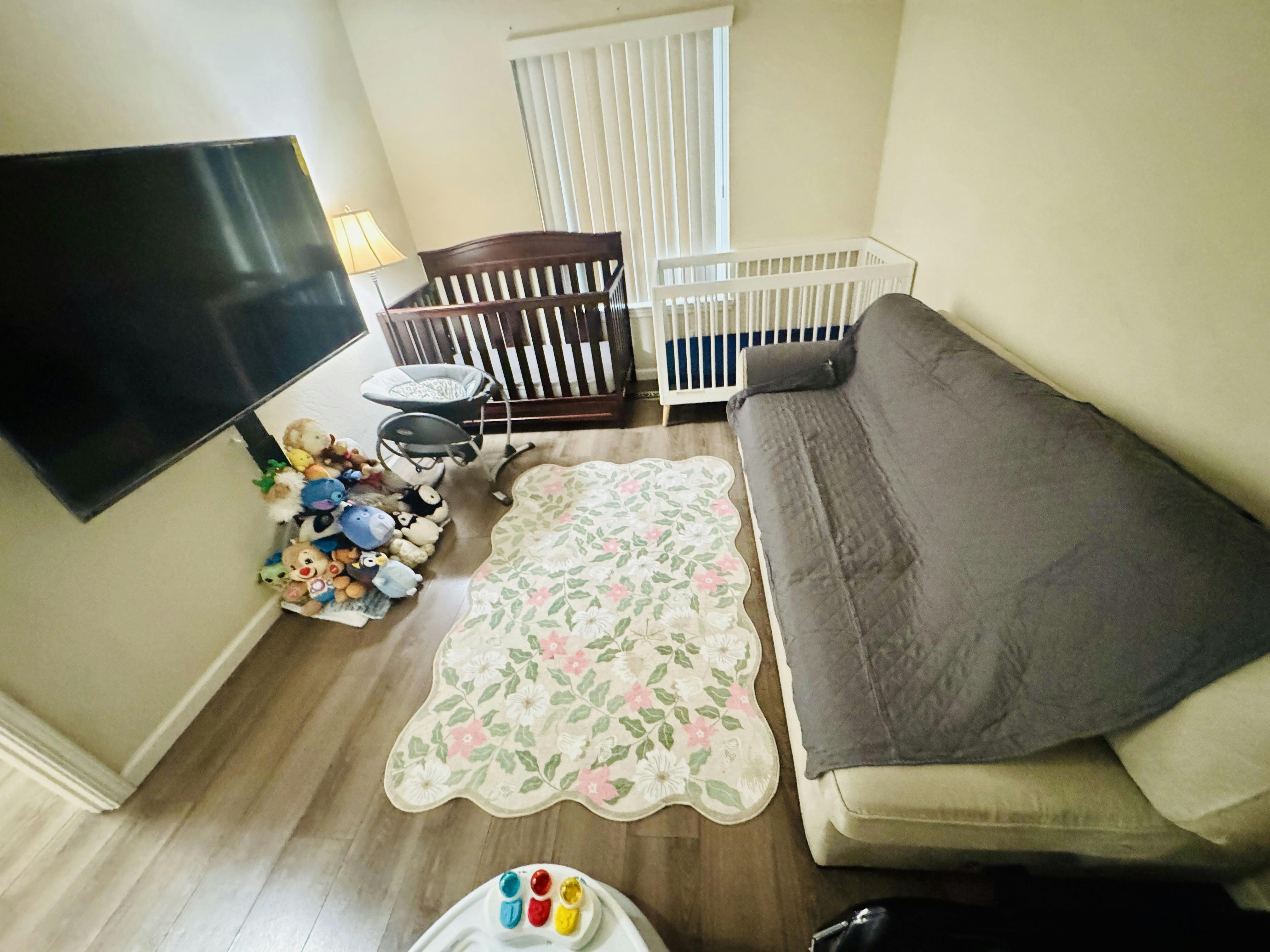 A room with two cribs, a sofa, a television, and a floral rug