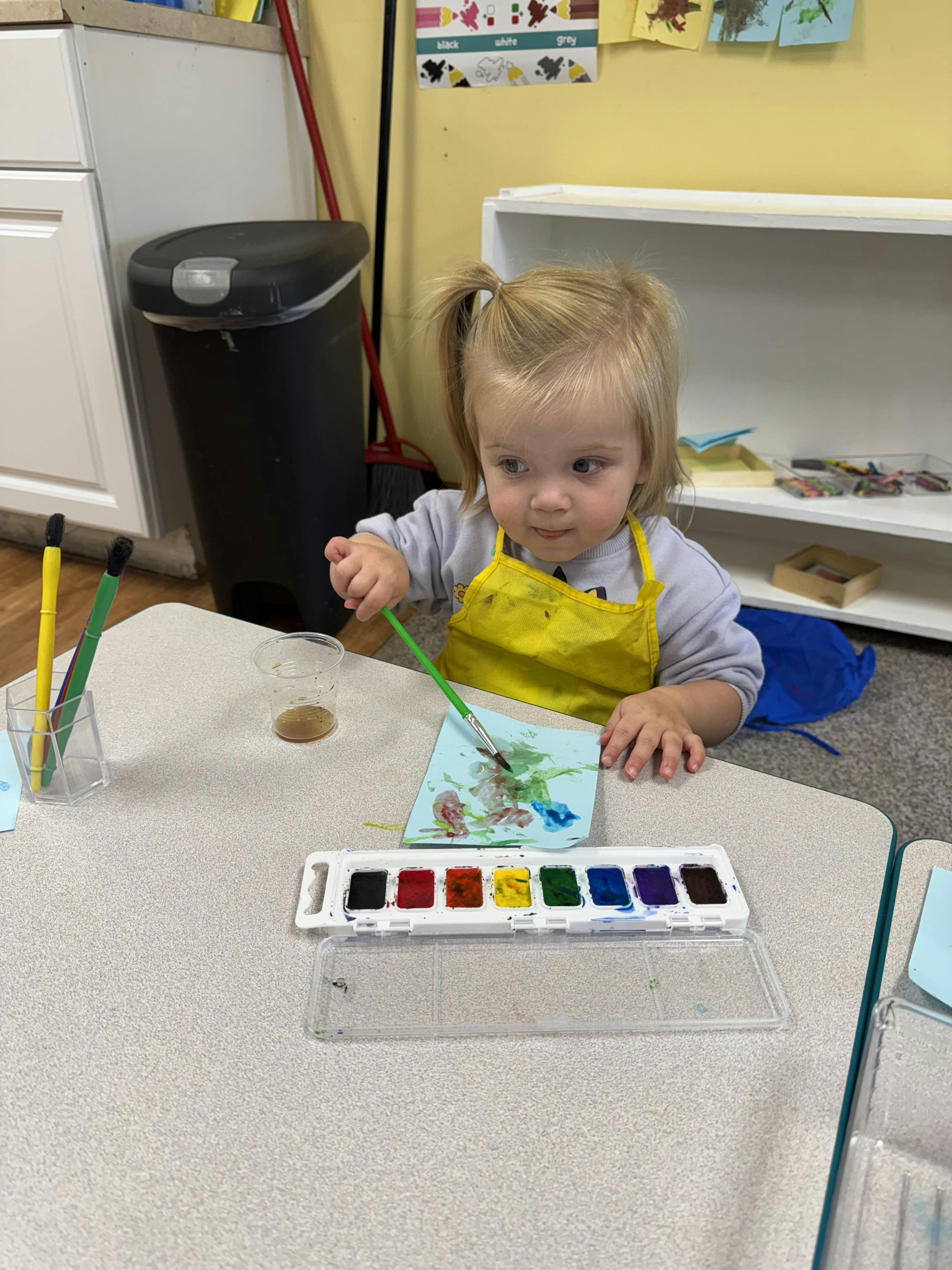 Child in a yellow apron painting with watercolors at a table