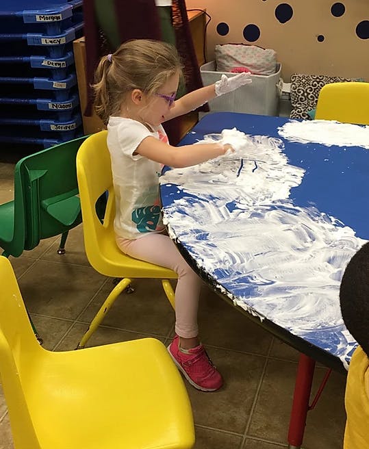 Child playing with shaving cream on a blue table in a classroom