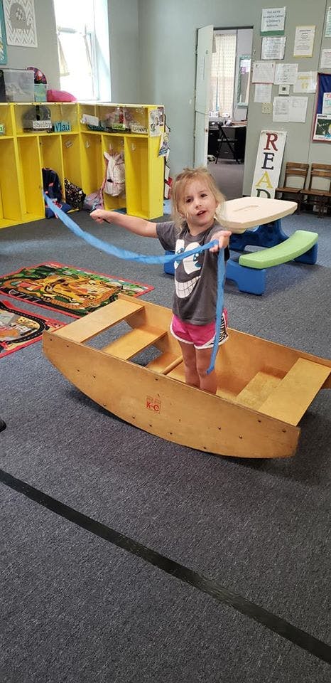 Child standing in a wooden rocking boat holding a blue ribbon