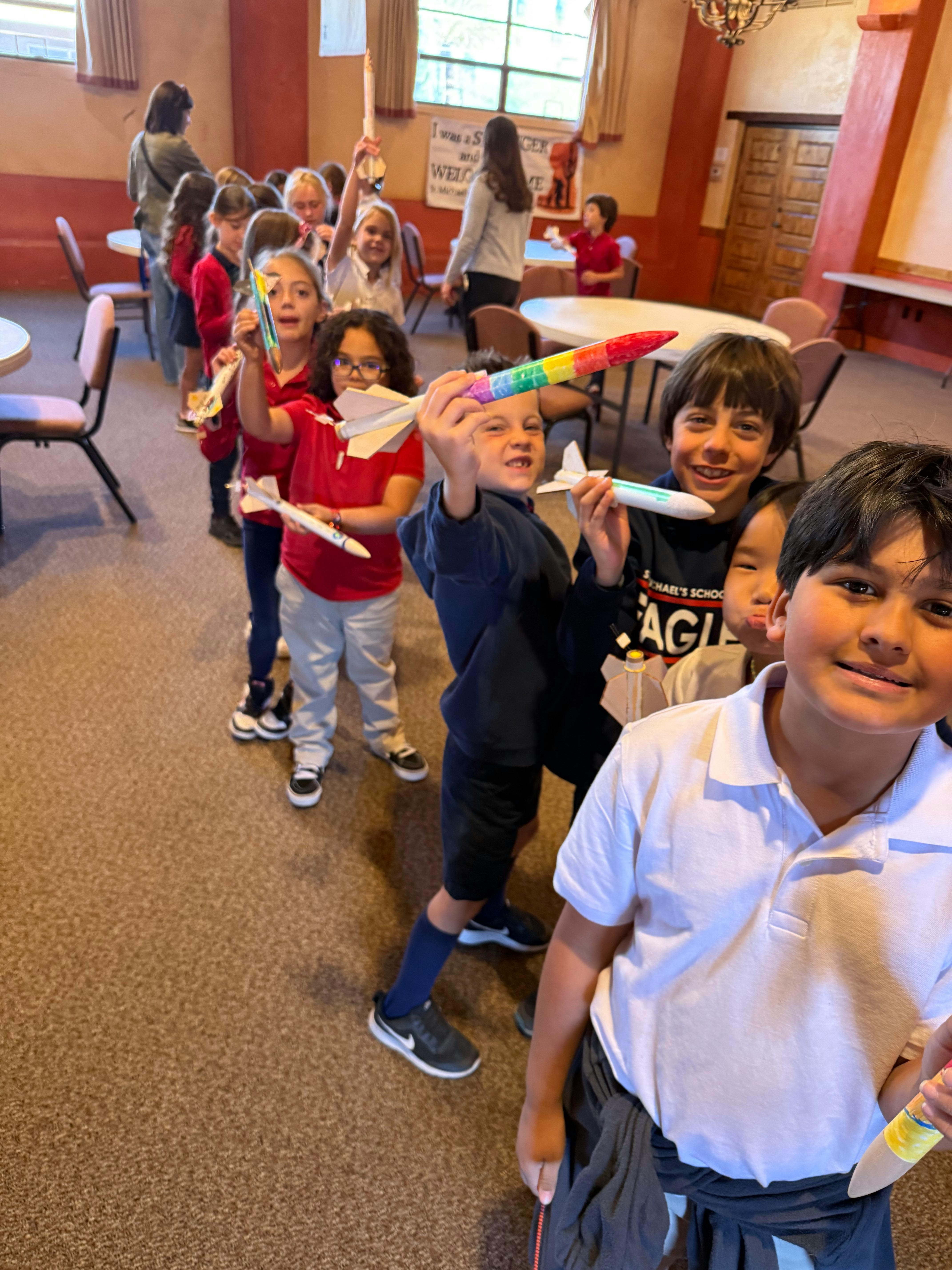 Children in a line holding up paper airplanes and foam rockets