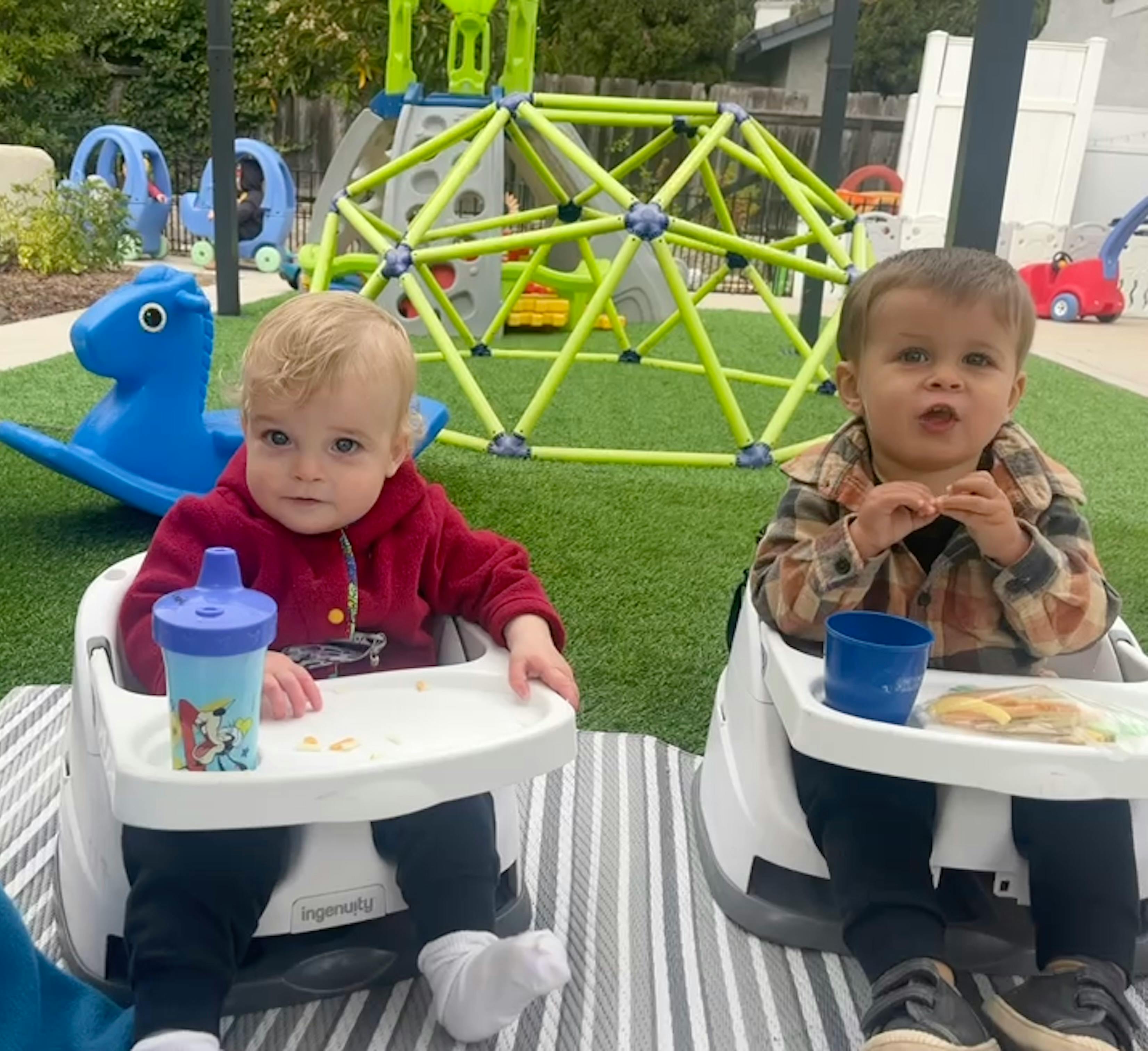 Two children in high chairs on a striped mat outdoors