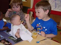 Child feeding a doll with a spoon, another child in background