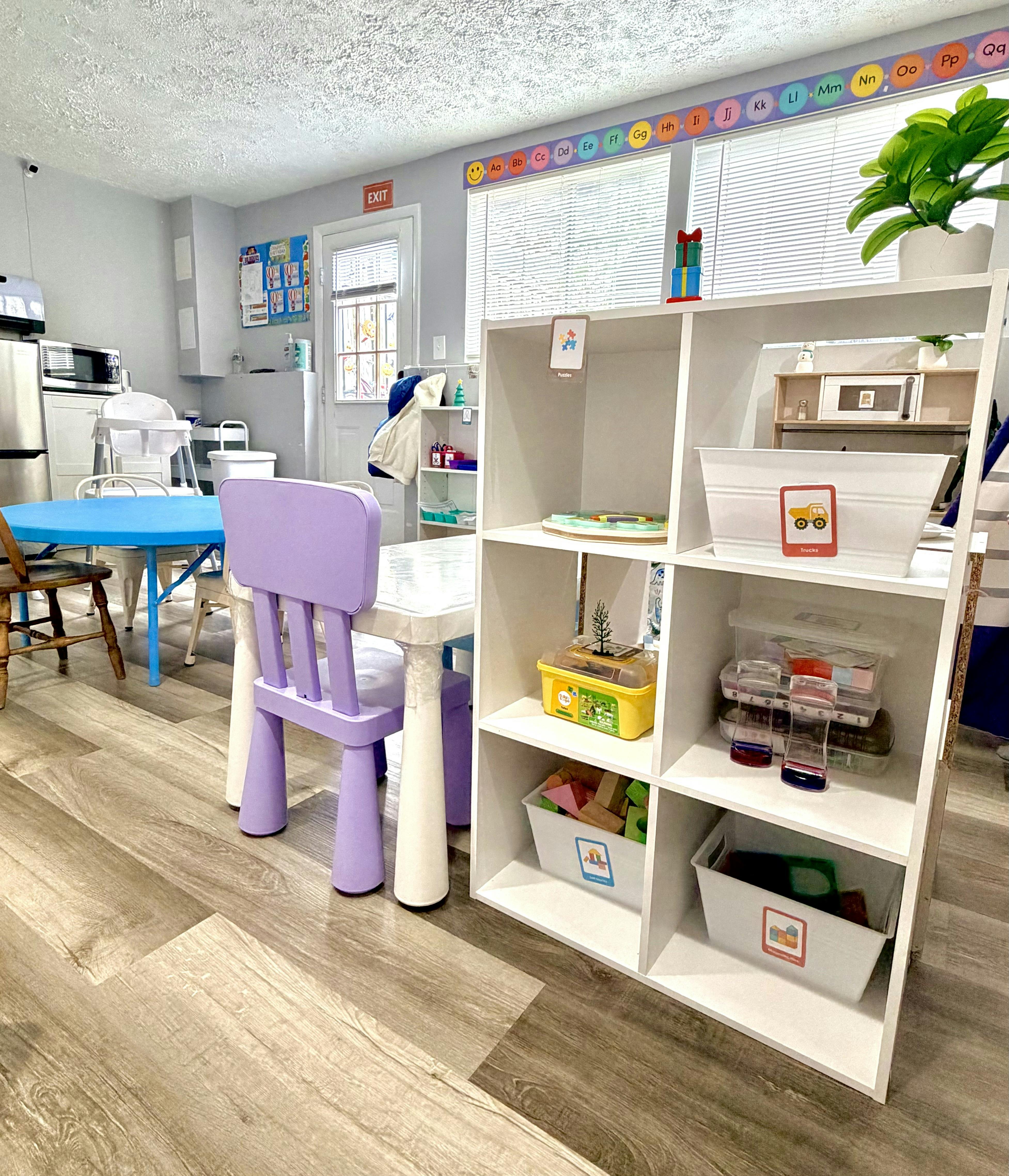 Classroom or daycare with small tables, chairs, shelving with toys, and an alphabet banner