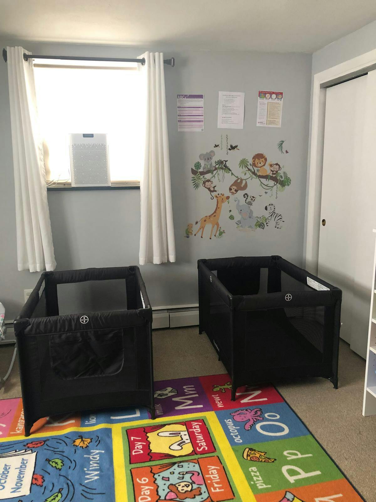 Two black portable cribs on an ABC rug in a room with a window and wall decals