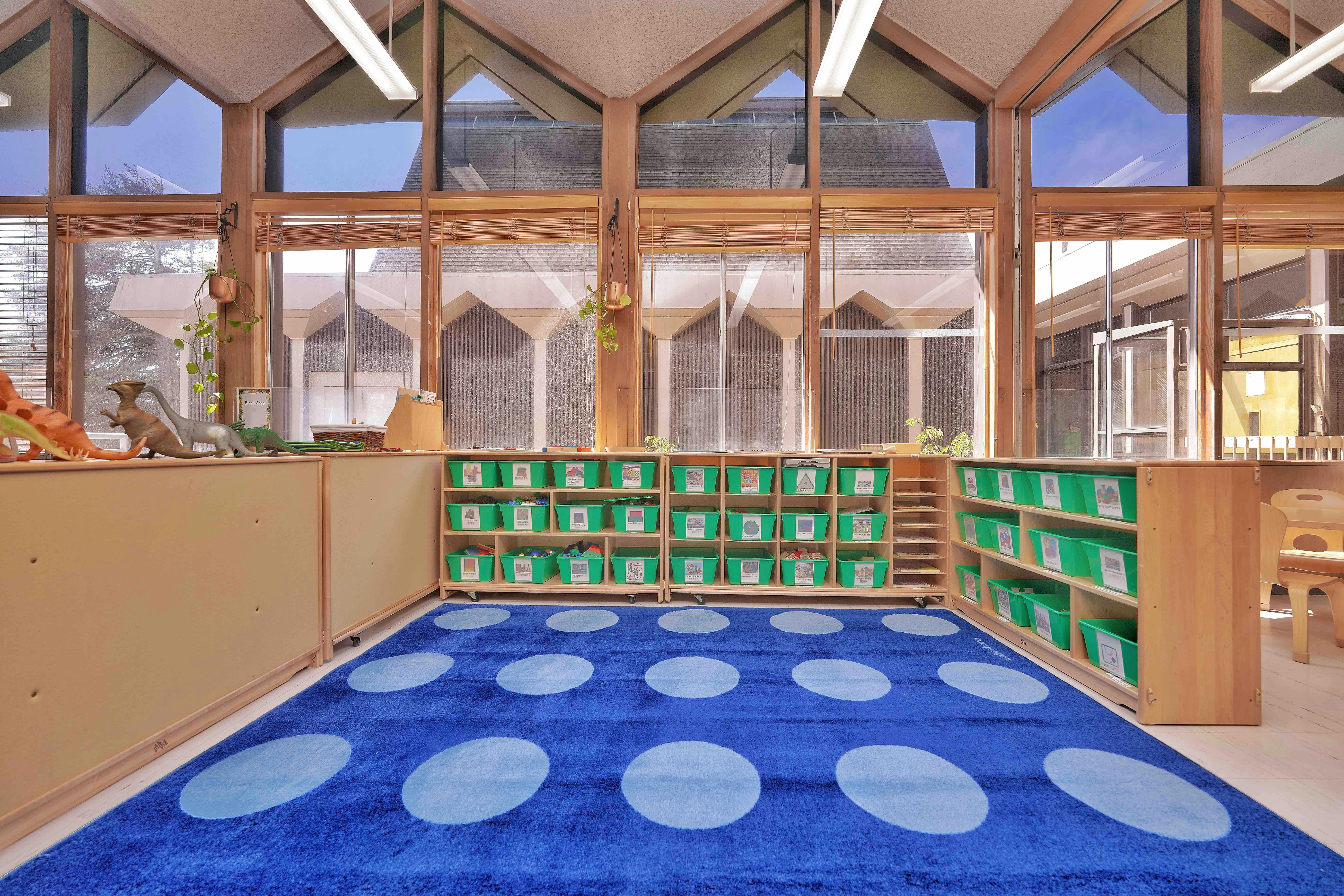 Classroom storage shelves with green bins and dinosaur toys on a blue dot rug