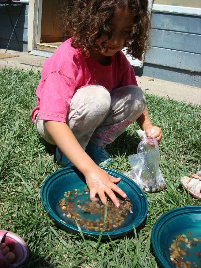 Child crouching in grass, hand in a bowl of water and pebbles