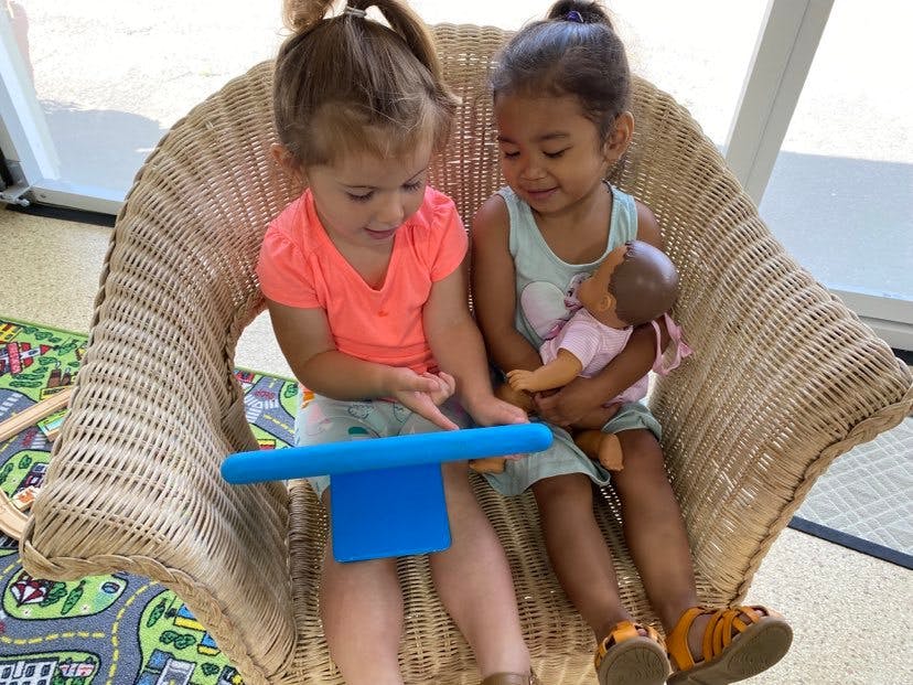 Two children sitting in a chair, one holding a doll and the other pointing at a tablet