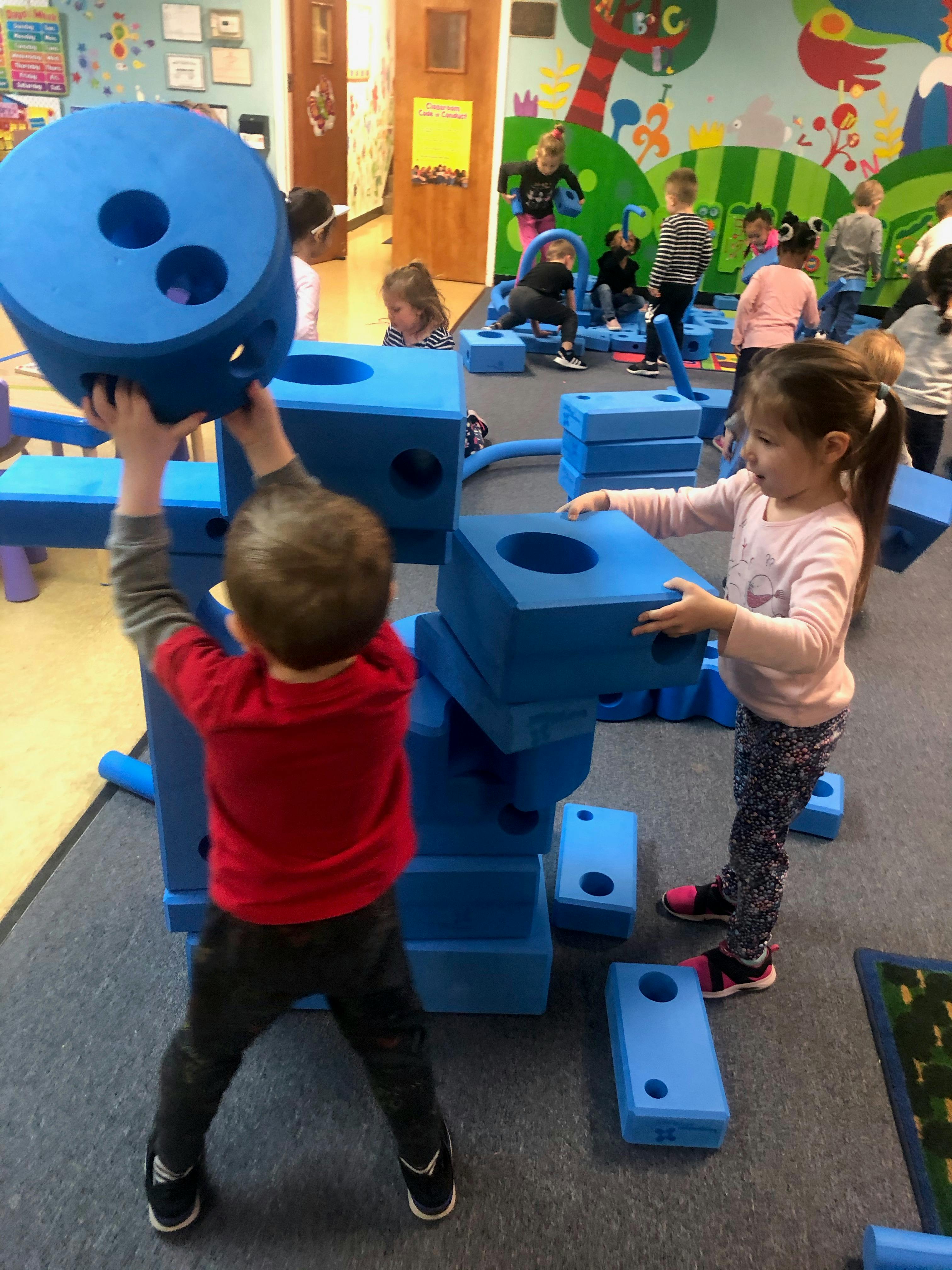 Two children playing with large blue building blocks in a classroom