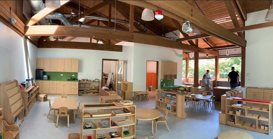 Classroom with wooden furniture, shelves, and small tables, kitchen area with green backsplash