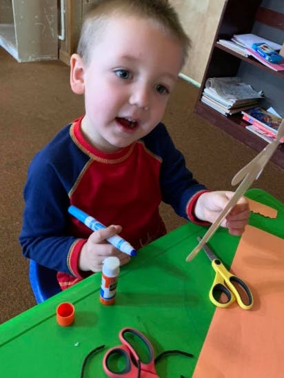Child holding a marker and a craft stick at a table with scissors and glue