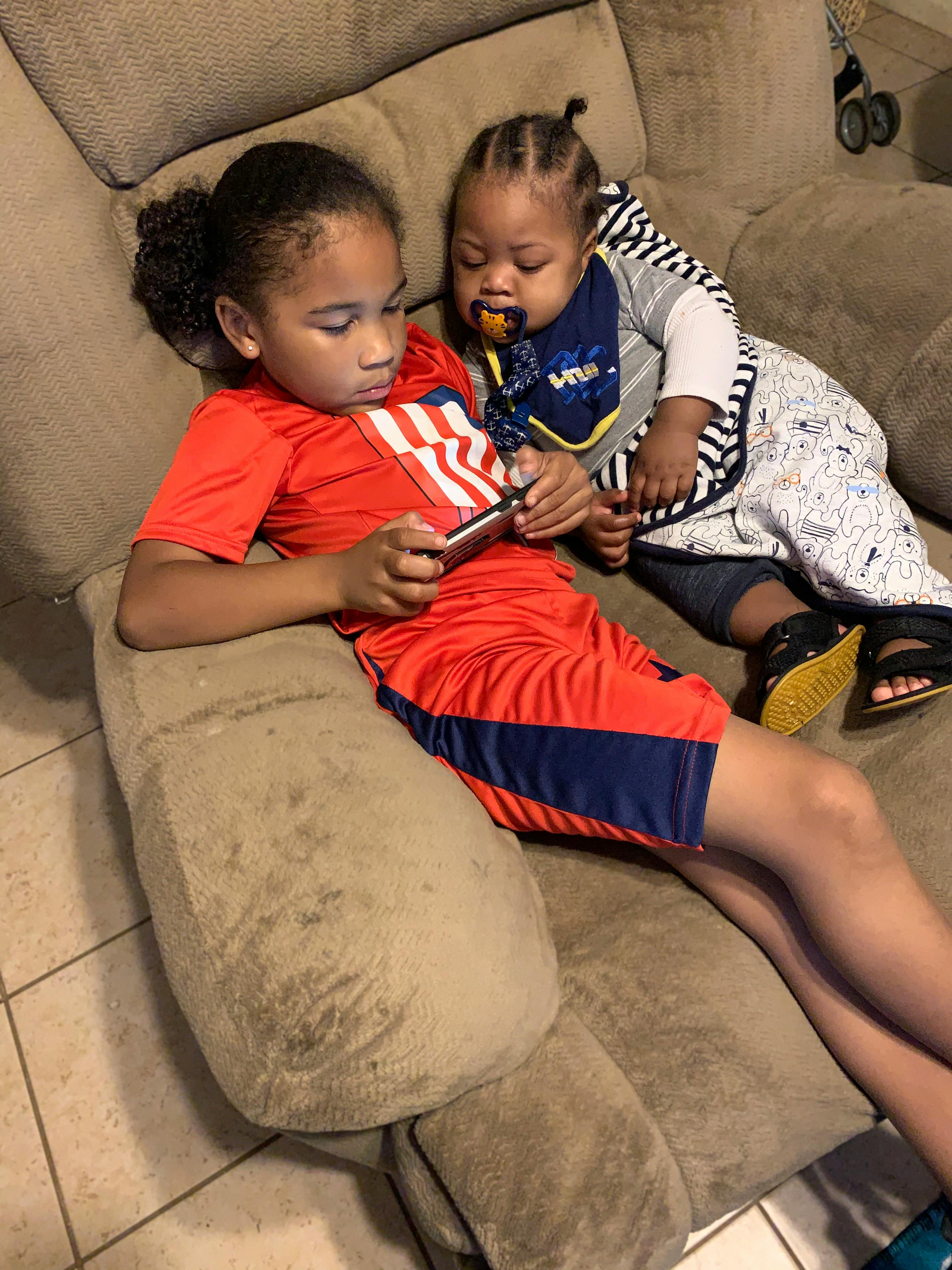 Child in red shirt holding a phone with a baby next to them on a couch